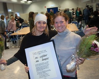 Två glada pristagare med diplom och blommor framför åhörare